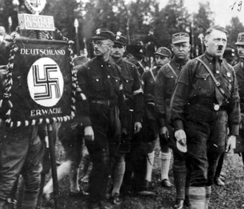 Auf dem Bild ist Adolf Hitler Er hat einen Schnurrbart. Neben ihm stehen andere Männer in Uniform.Links sieht man eine Fahne mit einem Kreuz. Das Kreuz nennt man Haken·kreuz. Das Haken·kreuz ist das Zeichen der Nazis.
