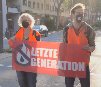 Das Foto zeigt eine Protestaktion der Letzten Generation in Heidelberg. Menschen stehen auf der Straße. Sie blockieren die Straße. Sie haben ein Plakat. Auf dem Plakat steht: Letzte Generation.